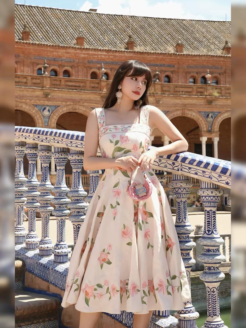 May include: A cream dress adorned with a floral design of pink flowers and green foliage. The dress features a square neckline and broad straps. A small pink purse is held. The backdrop showcases a building with arches and decorative columns.