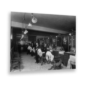 May include: A black and white photograph of a barbershop interior. The shop has multiple barber chairs with customers seated in them. The barbers are standing behind the chairs, waiting to cut hair. The shop is decorated with mirrors and lights.