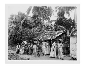 May include: A black and white photograph of a group of people standing in front of a simple, thatched-roof hut. The people are dressed in clothing typical of the time period, and some are holding children. The hut is surrounded by lush vegetation, including palm trees and banana plants.