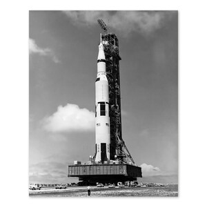 May include: A black and white photograph of a rocket on a launch pad. The rocket is tall and slender, with a pointed nose cone and a large, cylindrical body. The launch pad is a large, metal structure with a series of platforms and supports. The rocket is surrounded by a cloud of smoke and steam.