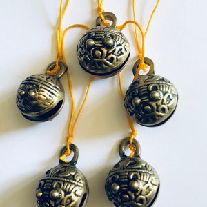 May include: Five antique bronze bells with intricate floral patterns, each suspended by a thin yellow cord. The bells have a tarnished finish, suggesting age and use. The arrangement is against a plain white background.