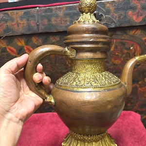 May include: A copper-coloured teapot with a decorative gold-coloured base, handle, and accents. The teapot has a bulbous body, a curved handle, and a lid with a floral finial. The teapot is likely an antique or vintage item.