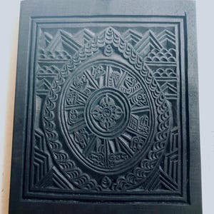 May include: A black wooden plaque with a carved design. The design features a circular pattern with intricate details and a central motif. The plaque has a small wooden handle at the top.
