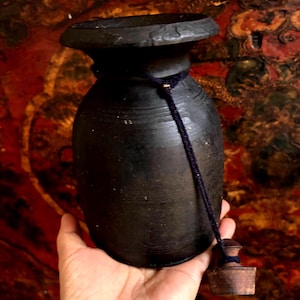 Puede incluir: Un jarrón de cerámica negro con un cordón azul oscuro y un tapón de madera. El jarrón tiene un cuello estrecho y una base ancha.