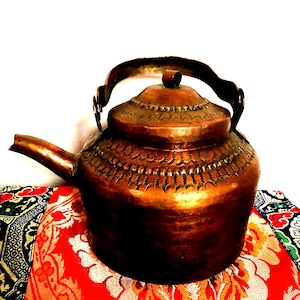 May include: An antique copper teapot with an ornate engraved design. The teapot has a rounded body and a lid with a small knob. The handle is made of copper and has a simple design.