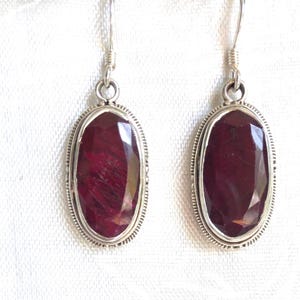 May include: A pair of oval-shaped ruby earrings. Each earring features a deep red gemstone set in a silver-colored metal frame with a decorative border. The earrings have a simple hook closure.