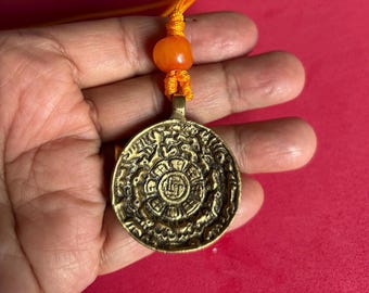 Puede incluir: Un colgante redondo de color oro antiguo con intrincados diseños en relieve. El colgante está suspendido de un cordón naranja con una cuenta naranja. El colgante se sostiene en una mano sobre un fondo rojo.