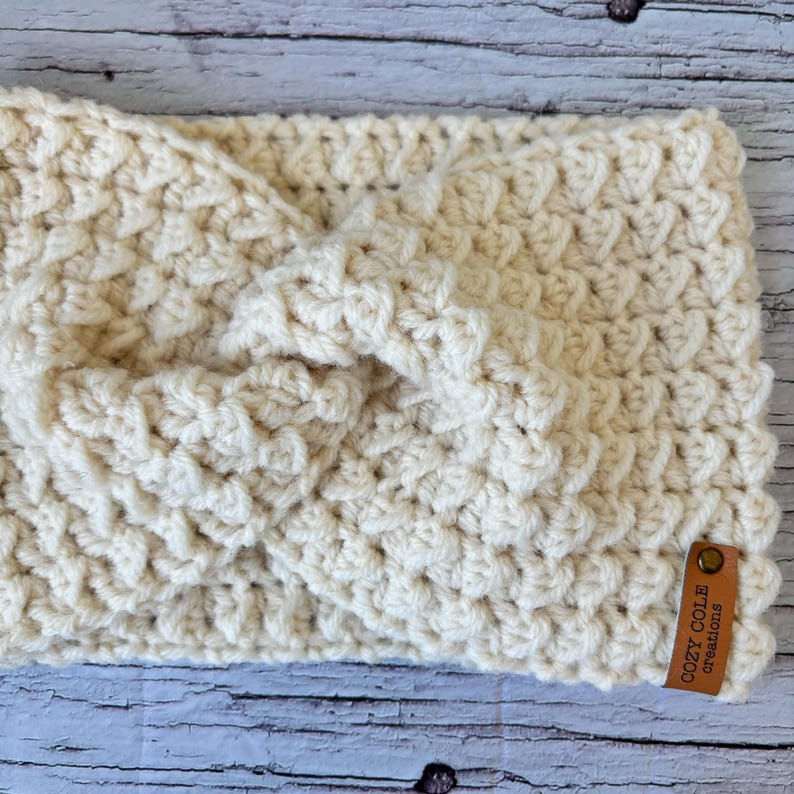 Op de afbeelding: Een cr&egrave;mekleurig gehaakt hoofdband met een geknoopt ontwerp. Het hoofdband heeft een bruin lederen label met de tekst "Cozy Cole Creations" erop.