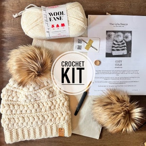 May include: Crochet kit for a beanie hat with a faux fur pom pom. The kit includes white yarn, a crochet hook, and a pattern for the "Cozy Cole Lyla Beanie".