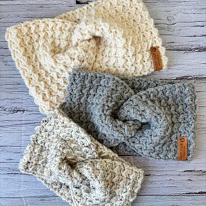Op de afbeelding: Drie gebreide hoofdbanden in verschillende kleuren en patronen. De hoofdbanden zijn gemaakt met een dik garen en hebben een geknoopt ontwerp. De hoofdbanden zijn voorzien van een bruin lederen label met de tekst "Cozy Coils Handmade".