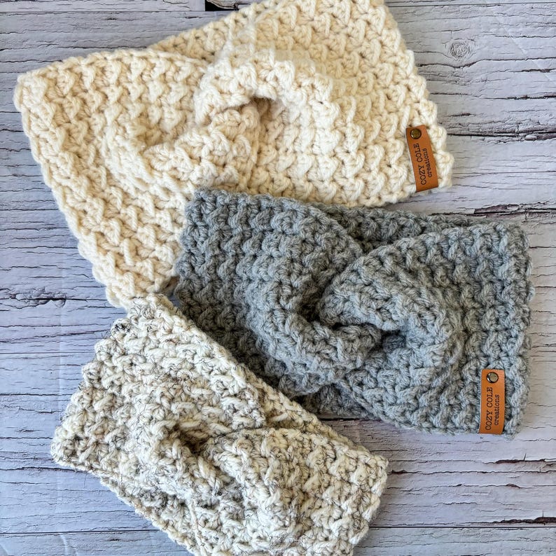 Op de afbeelding: Drie gebreide hoofdbanden in verschillende kleuren en patronen. De hoofdbanden zijn gemaakt met een dik garen en hebben een geknoopt ontwerp. De hoofdbanden zijn voorzien van een bruin lederen label met de tekst "Cozy Coils Handmade".