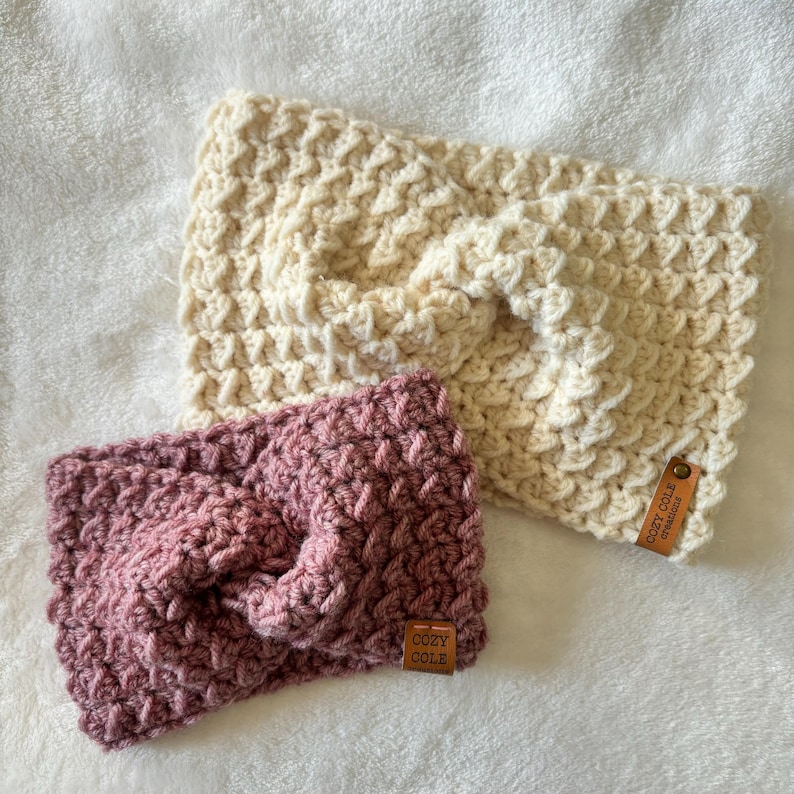 Op de afbeelding: Twee gebreide hoofdbanden, een in cr&egrave;mekleur en een in een stoffige roze kleur. Beide hoofdbanden hebben een geknoopt ontwerp en een bruin lederen label met de tekst "Cozy Cole" erop.