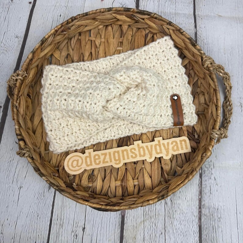 Op de afbeelding: Een cr&egrave;mekleurig gehaakt hoofdband met een geknoopt ontwerp. Het hoofdband wordt getoond in een geweven mand met een houten label waarop "@dezignsbydyan" staat.