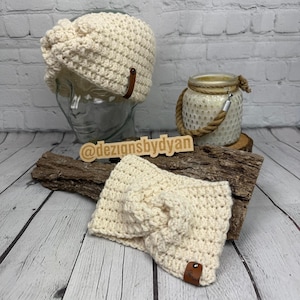 Op de afbeelding: Twee cr&egrave;mekleurige gehaakte haarbanden met een bruin lederen accent. De haarband links bevindt zich op een mannequinhoofd. De haarband rechts bevindt zich op een houten oppervlak. De tekst "@dezignsbydyan" is zichtbaar op een houten bord.