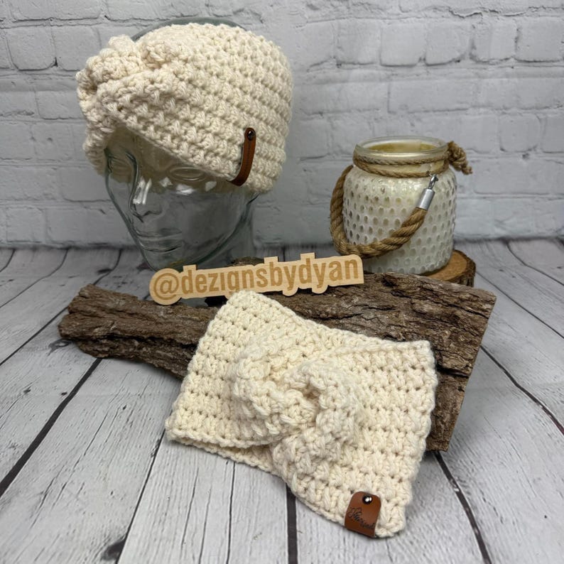 Op de afbeelding: Twee cr&egrave;mekleurige gehaakte haarbanden met een bruin lederen accent. De haarband links bevindt zich op een mannequinhoofd. De haarband rechts bevindt zich op een houten oppervlak. De tekst "@dezignsbydyan" is zichtbaar op een houten bord.