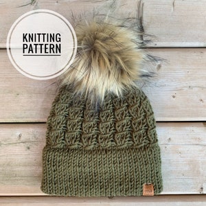 May include: An olive green knitted hat with a textured pattern and a faux fur pom-pom. The hat has a ribbed brim and a small leather tag that reads "COZY COLE". The image also includes the text "KNITTING PATTERN" in a white circle.
