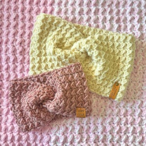 Op de afbeelding: Twee gebreide hoofdbanden, een in een lichtgele kleur en een in een stoffige roze kleur. Beide hoofdbanden hebben een geknoopt ontwerp en een klein lederen label met de tekst "Cozy Cole Handmade" erop.