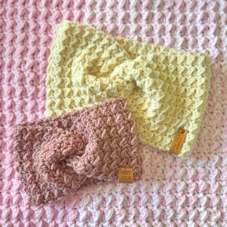 Op de afbeelding: Twee gebreide hoofdbanden, een in een lichtgele kleur en een in een stoffige roze kleur. Beide hoofdbanden hebben een geknoopt ontwerp en een klein lederen label met de tekst "Cozy Cole Handmade" erop.