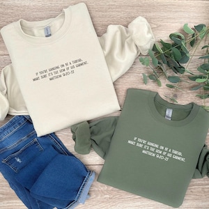 Könnte beinhalten: Zwei Sweatshirts mit dem gleichen Text: "Wenn du am seidenen Faden hängst, achte darauf, dass es der Saum seines Gewandes ist. Matthäus 9,20-22". Ein Sweatshirt ist beige und das andere grün.