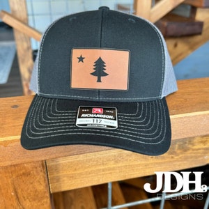 May include: Black and grey trucker hat with a brown leather patch featuring a pine tree and a star. The patch is labelled "Richardson" and "Original Trucker".