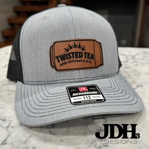 May include: Gray and black trucker hat with a brown leather patch that says "Twisted Tea and Double D's". The patch has a sun design and a can shape. The hat has a black mesh back and an adjustable strap with a Richardson 112 label.