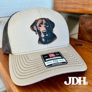 May include: A brown and black trucker hat with a brown leather patch of a black labrador retriever on the front. The hat has a black mesh back and a brown adjustable strap with the text "EST. 1970 RICHARDSON. ORIGINAL 112 TRUCKER ADJUSTABLE".