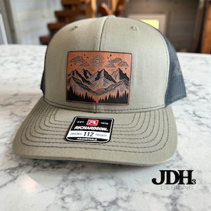 May include: A tan and black Richardson 112 trucker hat with a mountain and sun patch. The patch features a detailed illustration of mountains, trees, and a sun. The hat has a curved brim and an adjustable snap closure.