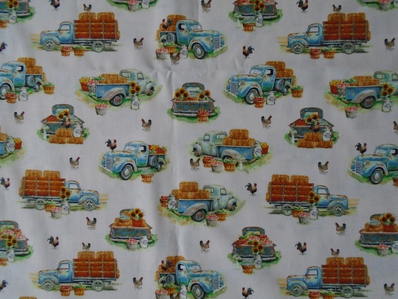 May include: A white fabric with a repeating pattern of blue vintage pickup trucks carrying hay bales, produce, and sunflowers. The trucks are surrounded by chickens and baskets of produce.