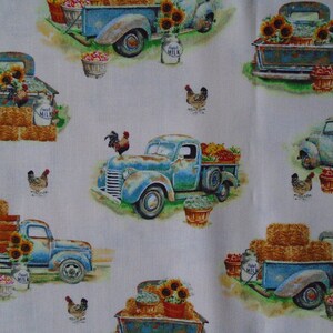 May include: A white fabric with a repeating pattern of blue vintage pickup trucks carrying hay bales, sunflowers, and produce. The trucks have a rustic, weathered look and are decorated with milk jugs and roosters.