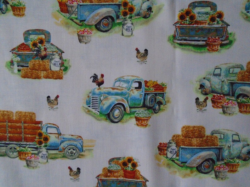 May include: A white fabric with a repeating pattern of blue vintage pickup trucks carrying hay bales, sunflowers, and produce. The trucks have a rustic, weathered look and are decorated with milk jugs and roosters.