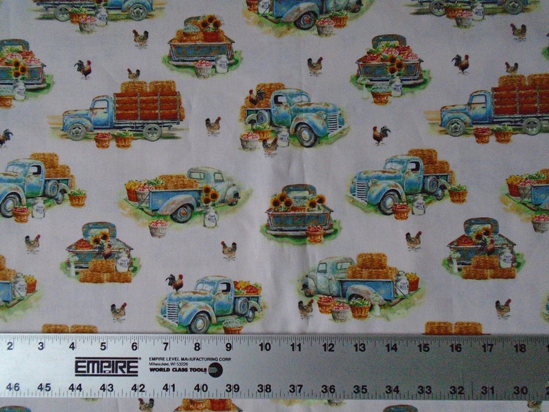 May include: A white fabric with a repeating pattern of blue pickup trucks carrying hay bales, sunflowers, and baskets of produce. The trucks are on a green grassy field with chickens and a ruler in the bottom of the image.