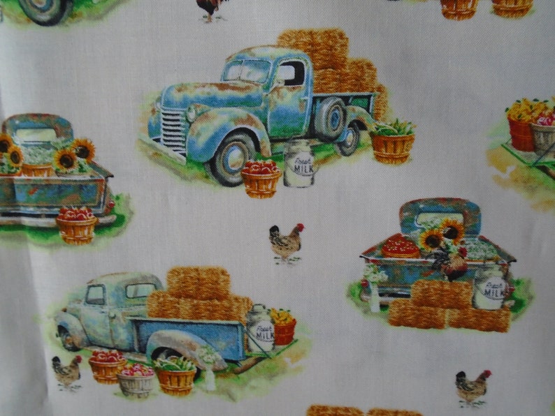 May include: A white fabric with a repeating pattern of blue vintage trucks, hay bales, sunflowers, chickens, and milk jugs. The trucks are carrying baskets of fruit and vegetables. The text "Good Milk" is printed on the milk jugs.