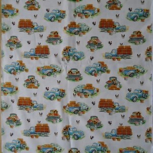 May include: White fabric with a repeating pattern of blue pickup trucks carrying hay bales. The trucks are surrounded by sunflowers, chickens, and baskets of produce.