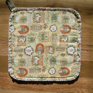 May include: Square beige pot holder with a repeating pattern of Tuscan-themed illustrations in orange, green, and brown. The design includes text such as "Tuscany" and "Ciao Bella". The edges are trimmed with a ruffled border and a hanging loop.