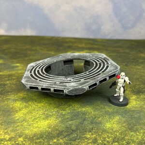 May include: A gray, circular, multi-level platform with a large hole in the center. The platform is made of plastic and has a textured surface. It is sitting on a green grassy surface.