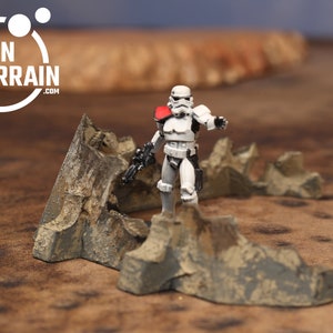 May include: A white stormtrooper action figure stands on a grey and brown rocky terrain model. The terrain model is made of plastic and is designed for tabletop gaming. The model is a realistic representation of a rocky landscape.