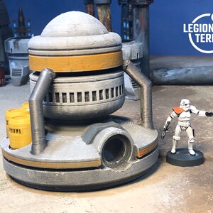 May include: A gray and yellow 3D printed model of a futuristic industrial building with a round top and a large pipe on the side. The building is sitting on a sandy surface and a white action figure is standing in front of it.