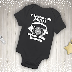 May include: Black baby onesie with white text that reads "I Listen To Metal With My Daddy". The onesie has a graphic of a microphone and headphones.