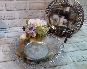 May include: Two silver decorative plates with intricate designs. The top plate has a larger diameter and is on a stand. The bottom plate is smaller and has a floral design around the edge. A bouquet of artificial flowers is on top of the smaller plate.