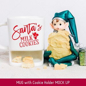 Puede incluir: Taza de cer&aacute;mica blanca con un soporte para galletas. La taza tiene texto rojo que dice "Santa's Milk and Cookies". Un mu&ntilde;eco de duende verde est&aacute; sosteniendo una galleta y sentado junto a la taza.