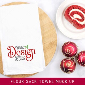 May include: A white flour sack towel mockup with the text "Your Design Here" in red and green. The towel is on a wooden cutting board with red and gold Christmas ornaments and a slice of red velvet cake roll on a white plate.