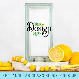 May include: A rectangular glass block mockup with a white background and the text "Your Design Here" in black. The glass block is sitting on a wooden surface with lemons and a yellow and white beaded garland.