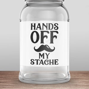 以下が含まれることがあります： 銀色の蓋と白いラベルが付いた透明なガラス瓶。ラベルには黒い口ひげのグラフィックと共に「Hands Off My Stache」と書かれている。