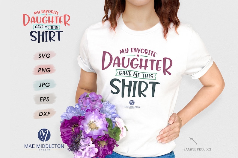 May include: White t-shirt with the text "My Favorite Daughter Gave Me This Shirt" in a colorful, handwritten font. The shirt is being worn by a person with brown hair and a bouquet of purple flowers in their hand.