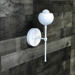 May include: White wall sconce with a round, white shade and a long, straight arm. The sconce is mounted on a white wall.