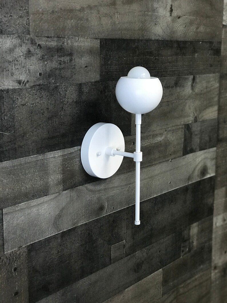 May include: White wall sconce with a round, white shade and a long, straight arm. The sconce is mounted on a white wall.