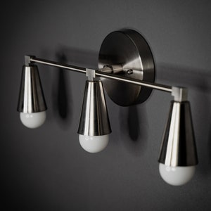 May include: A silver three-light wall sconce with a round backplate and three cone-shaped shades.