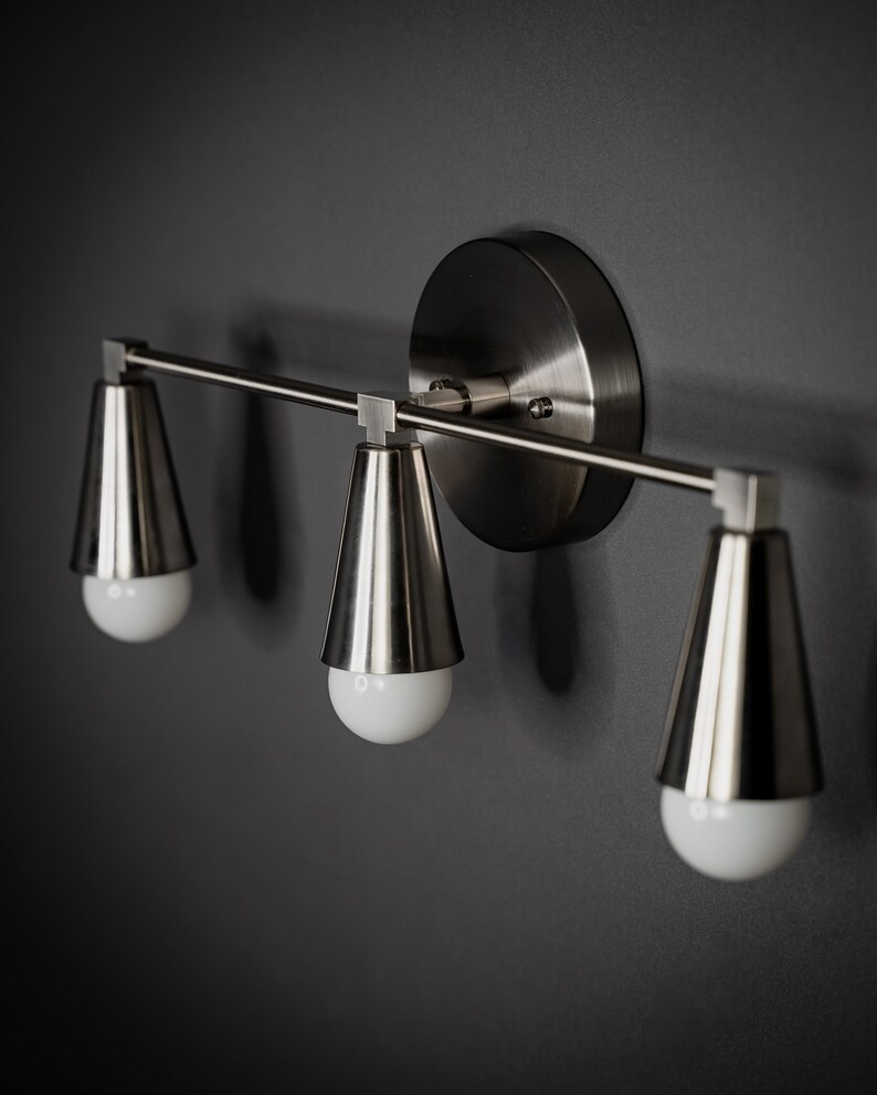 May include: A silver three-light wall sconce with a round backplate and three cone-shaped shades.