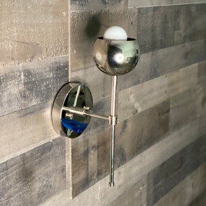 May include: A silver wall sconce with a round, mirrored base and a round, silver shade. The shade has a white light bulb inside.