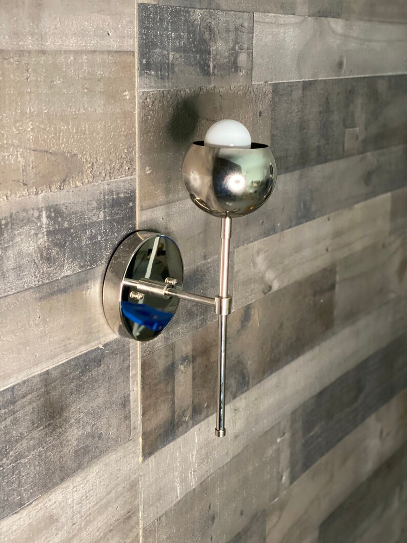 May include: A silver wall sconce with a round, mirrored base and a round, silver shade. The shade has a white light bulb inside.